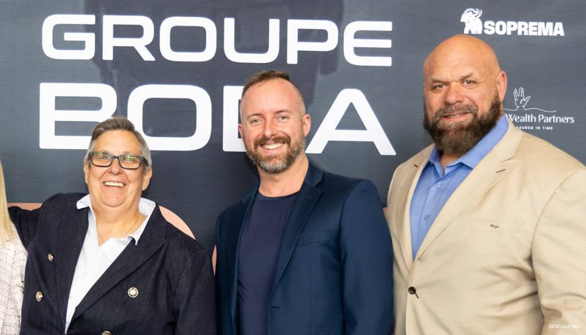 Photo de groupe à l'événement EQCO 2025. De gauche à droite : France Goyet (souriante avec lunettes et veston noir), Philippe Campagna-Vézina (souriant en veston bleu marin) et l'homme fort Hugo Girard (barbu en veston beige), posant devant un mur de commanditaires affichant Groupe Boda et Soprema.