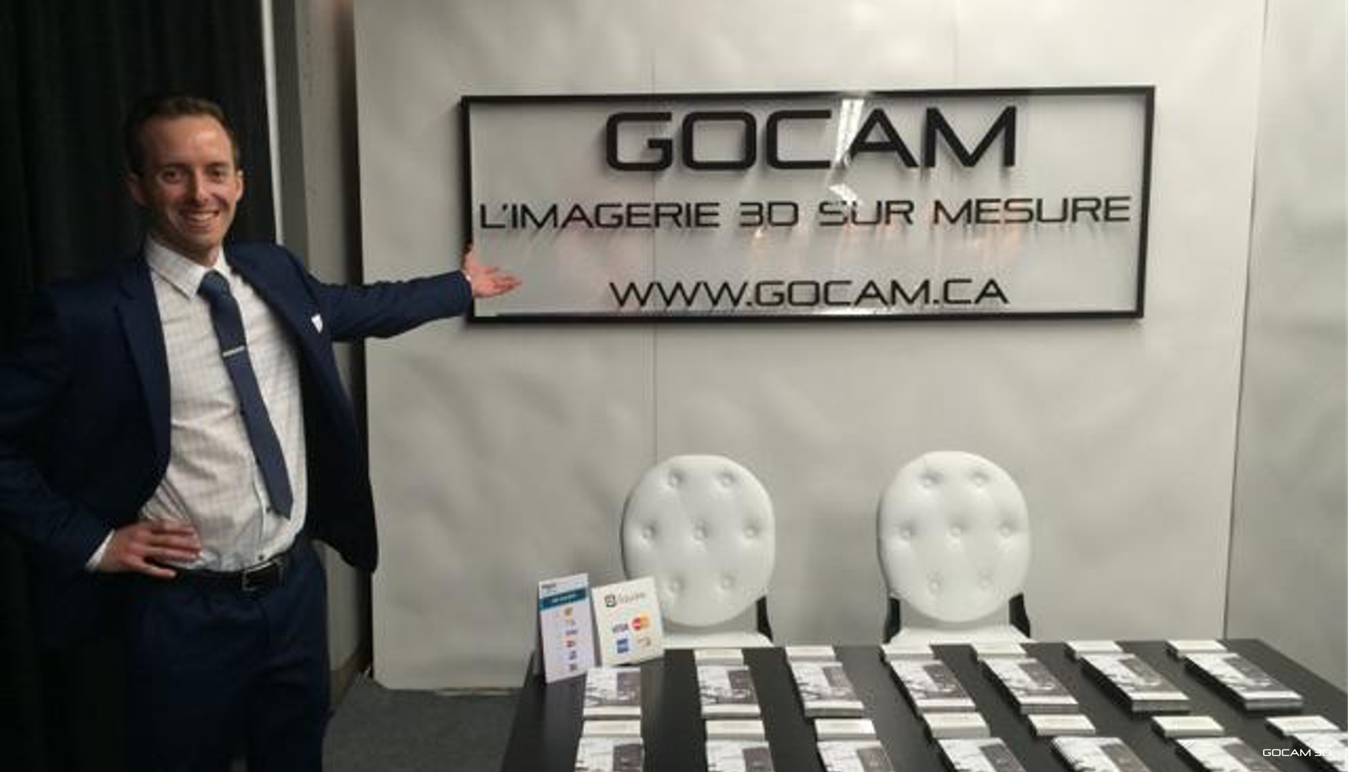 Philippe Campagna-Vézina en complet bleu souriant et présentant le kiosque officiel de GOCAM 3D lors du Salon ExpoHabitation de février 2016. À l'arrière-plan, une grande enseigne affiche le logo GOCAM, le slogan 'L'imagerie 3D sur mesure' et l'adresse du site web www.gocam.ca.