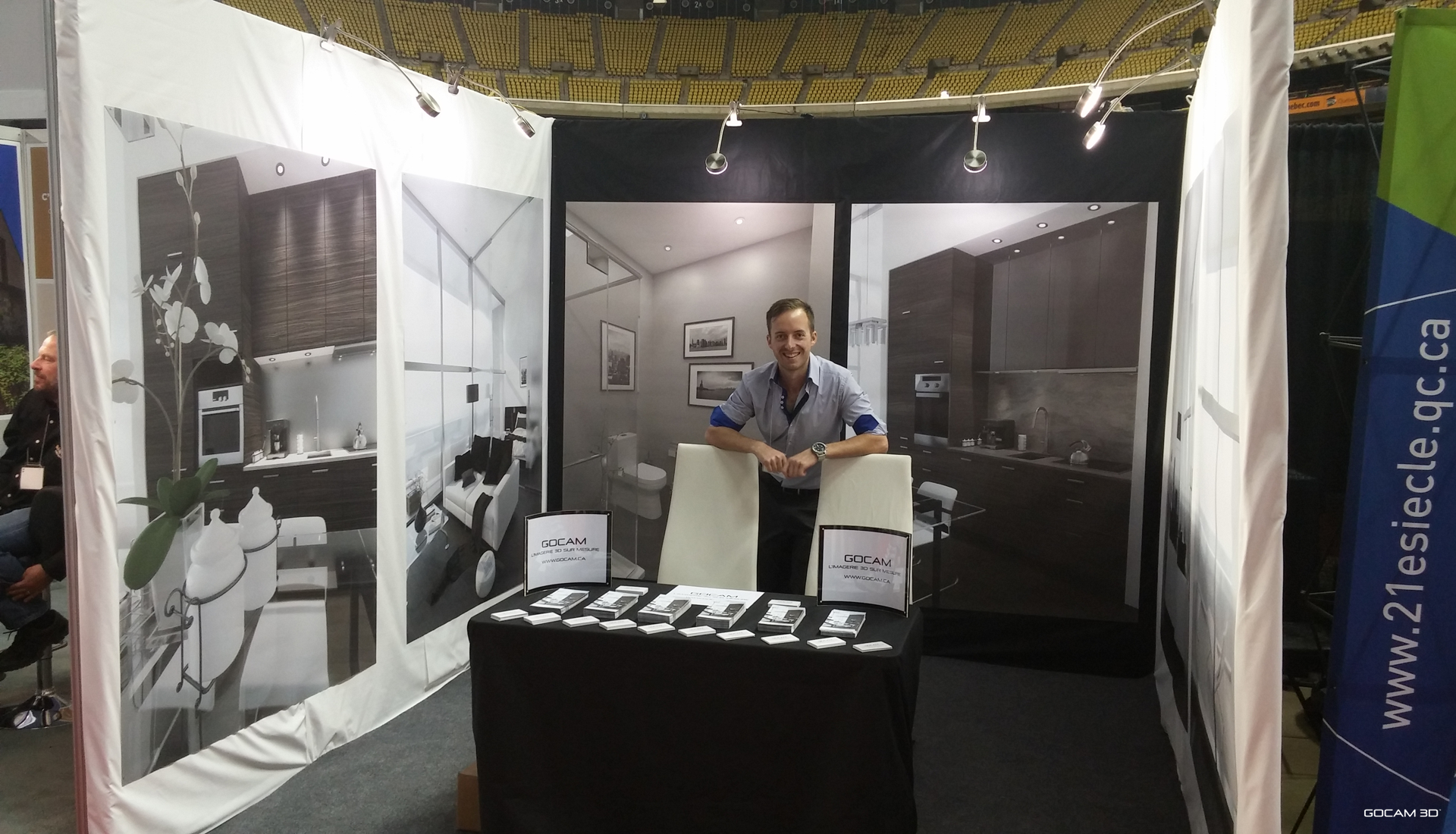 Philippe Campagna-Vézina souriant au kiosque d'exposition de GOCAM 3D lors du Salon National de l'Habitation de Montréal en octobre 2015, entouré de grands panneaux affichant des modélisations et perspectives 3D de cuisines, salons et salles de bain.