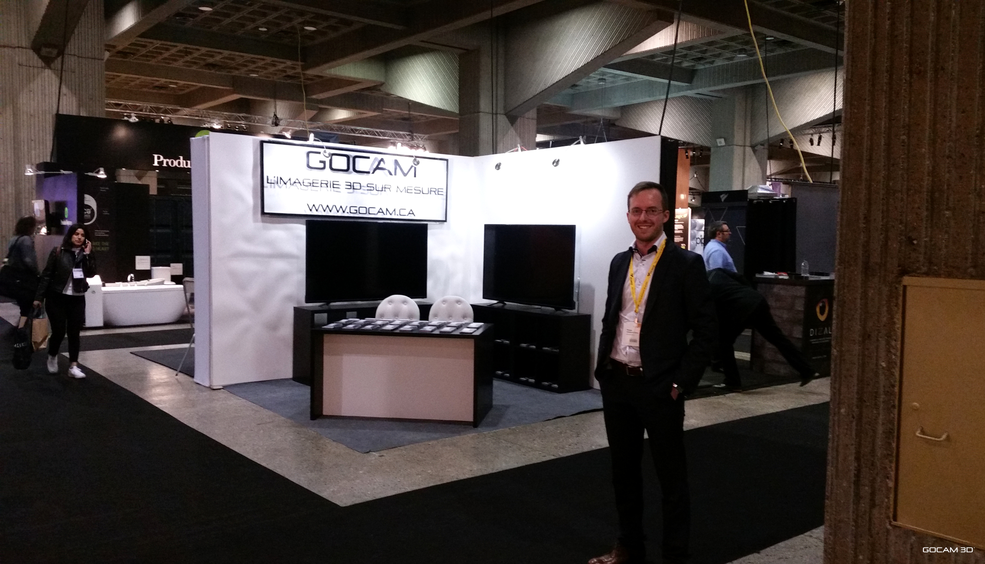 Philippe Campagna-Vézina debout devant le grand stand d'exposition de GOCAM 3D au SIDIM (Salon du Design) en mai 2017 à la Place Bonaventure de Montréal. Le kiosque moderne comprend une grande enseigne lumineuse, des comptoirs de présentation et deux grands écrans de télévision noirs.
