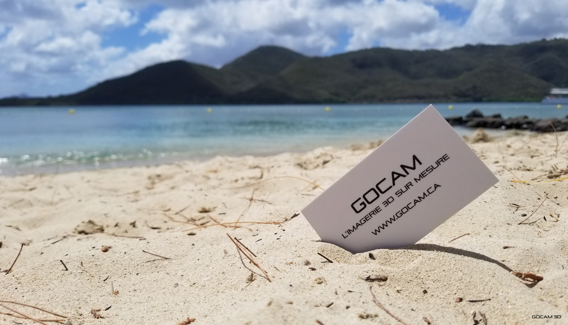 Une carte d'affaires blanche de GOCAM avec le slogan 'L'imagerie 3D sur mesure' plantée dans le sable chaud d'une plage en Martinique. À l'arrière-plan, une mer turquoise calme, des montagnes verdoyantes sous un ciel bleu parsemé de nuages blancs lors d'un voyage en 2019.